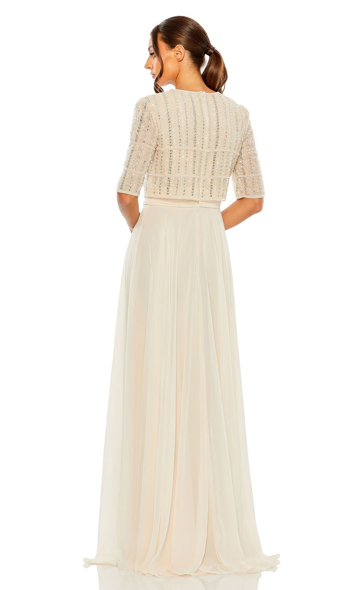 Woman standing with back to camera, wearing the Mac Duggal 9180 short sleeve long formal dress—a cream floor-length gown with a beaded crop top bodice and flowing A-line skirt.